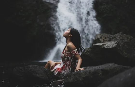 Frau sitzt entspannt auf einem Felsen vor einem Wasserfall